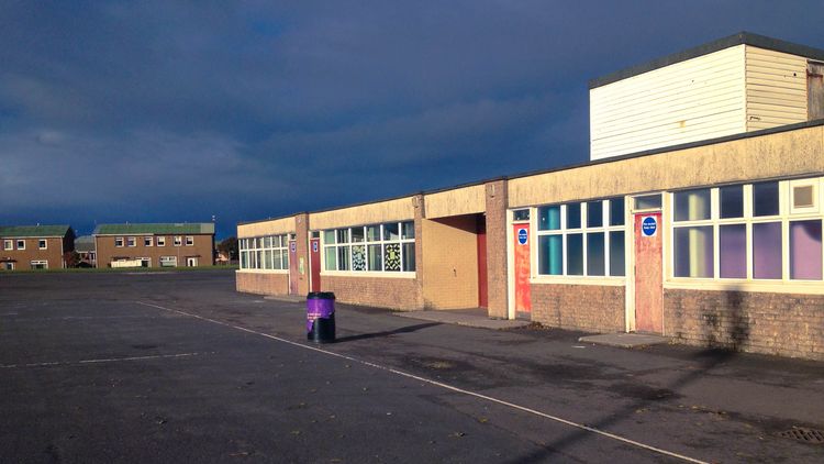 A primary school building in Prestwick, Scotland, in bright sunshine with dark, moody clouds above. 